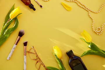 Yellow feminine flat lay: makeup brushes, glasses, oil, serum dropper, pearls. Yellow tulips and shadows. Beauty card concept. Copy space