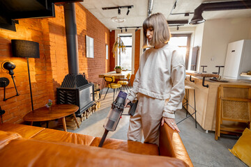 Young woman vacuuming leather couch with wireless handheld vacuum cleaner in modern living room at home. Housewife doing some housework © rh2010