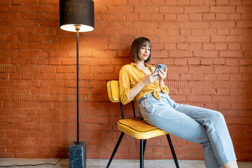 Young stylish woman using smart phone while sitting on chair near floor lamp on brick wall...