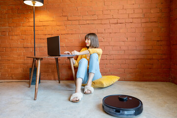 Young woman work on laptop computer, sitting on floor while robotic vacuum cleaner cleaning floor at home. Concept of smart home gadgets and work from home