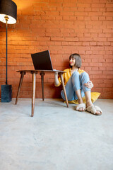 Young woman works on laptop computer while sitting relaxed by the coffee table on brick wall...
