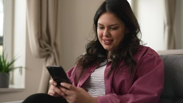 Portrait of young overweight woman using smartphone and sitting on sofa in home room spbd. 4k Closeup view of beautiful oversized female holds phone in hand and looks with smile, chats on social media