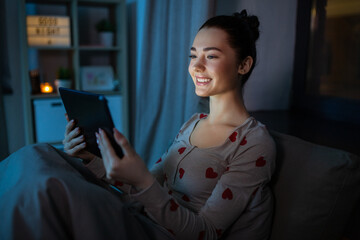 technology, bedtime and people concept - happy smiling teenage girl tablet pc computer sitting in bed at home at night