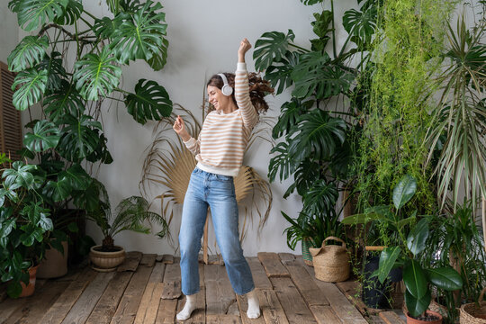 Carefree young woman with long hair in wireless headphones listening to music with pleasure, enjoying the moment, relaxing dancing on wooden floor in cozy home garden with monstera and tropical plants