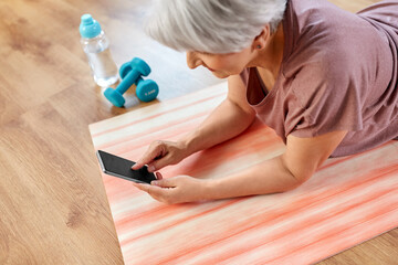 Obraz na płótnie Canvas sport, fitness and healthy lifestyle concept - smiling senior woman with smartphone exercising on mat at home