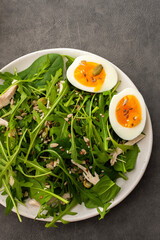 Delicious light salad with arugula and spinach, boiled eggs and chicken breast sprinkled with various healthy seeds in a plate on a gray background, top view
