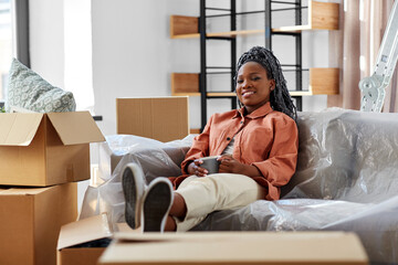moving, people and real estate concept - happy smiling woman with boxes sitting on sofa with cup of coffee at new home