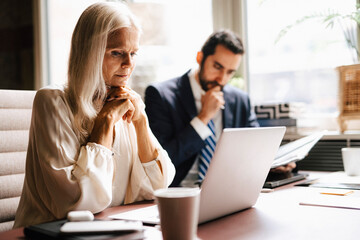 Mature businesswoman staring at laptop by businessman at desk in office