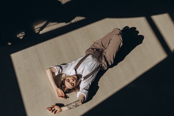 Portrait of a fashionable retro girl in a white shirt, suspenders and brown wide leg pants, sitting on the floor in the sun in the room
