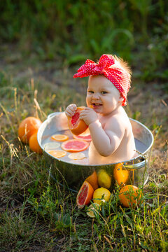 charming little girl in a red bandage is sitting in an iron tub with oranges and grapefruits in the garden and eating a slice of orange. outdoor water treatments. space for text. High quality photo