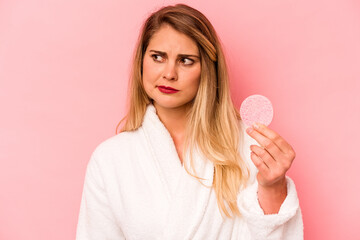 Young caucasian woman holding facial sponge isolated on pink background confused, feels doubtful and unsure.
