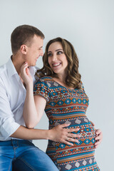 Happy pregnant couple on the grey background  is hugging and touching belly. Man in white shirt and pregnant woman in colorful ethnic dress