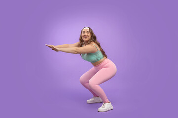 Fototapeta na wymiar Happy young pretty european plus size female in sportswear squats, isolated on purple background