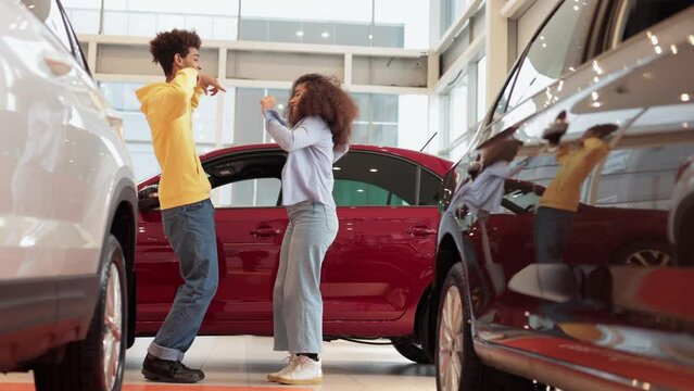 Young couple is dancing and having fun while shopping in car dealership office spbi. Beautiful European woman, man dance actively and look with smiles, pose for camera and stand on background of new