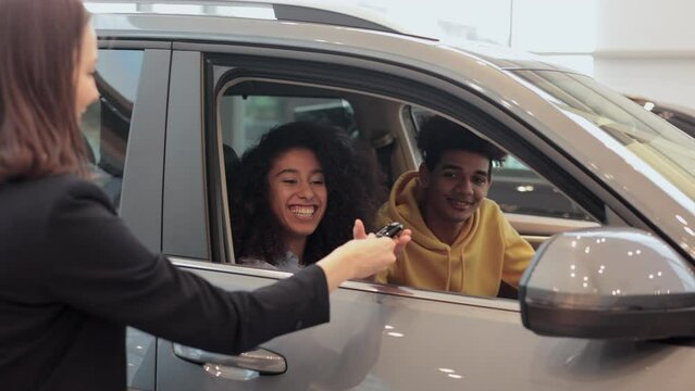 Young couple is sitting inside new car and taking keys from seller in company spbi. Close view of happy people customers sit in auto and look with smiles, get keys and talk to female manager at light
