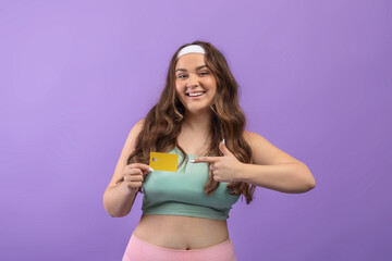 Cheerful young european plus size lady in sportswear point finger at credit card