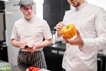Close-up of chef in uniform holding fresh pepper in his hands and talking to his assistant during...