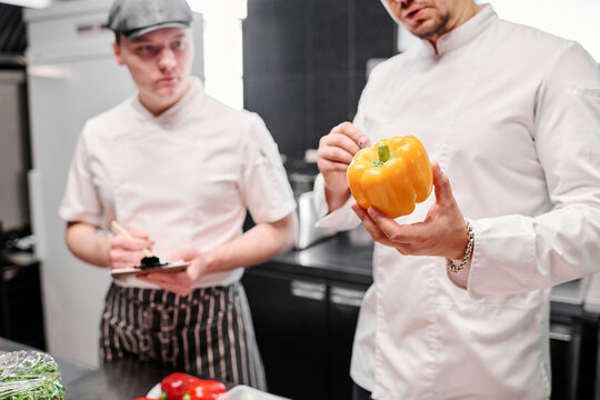 Close-up of chef in uniform holding fresh pepper in his hands and talking to his assistant during cooking