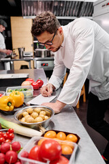 Young chef making a menu at table with food ingredients during his work in kitchen of restaurant