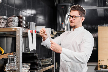 Young chef in uniform reading order of guest and starting to cook in kitchen