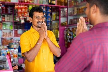 Smiling groceries merchant greeting by doing namaste to customer at kirana shop - concept of indian...