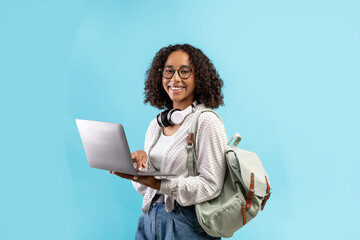 Cheerful young black woman with backpack and headphones holding laptop pc over blue studio...