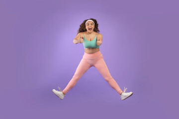 Satisfied excited young european plus size woman in sportswear jump, freeze in air, show thumbs up
