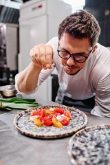 Young chef in eyeglasses adding spices in vegetable salad on plate while cooking at table