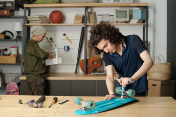 Contemporary teenage boy with wrench fixing wheels to blue skaeboard while bending over table...