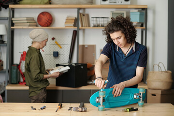 Contemporary teenager with handtool fixing wheels of skateboard by wooden table or workbench...