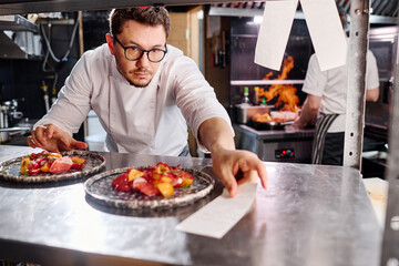 Young chef in eyeglasses putting list with order and prepared dishes on counter for delivery