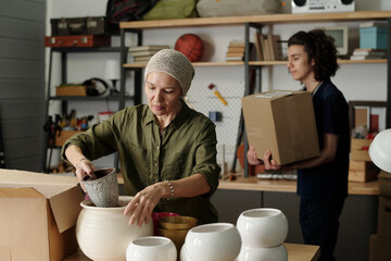 Female grandparent packing or unpacking boxes with various flowerpots while standing by table in...