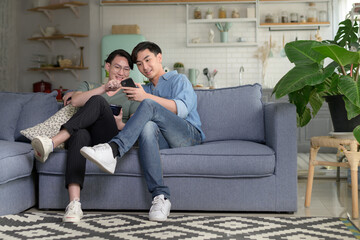 Obraz premium Young smiling gay couple using smartphone and relaxing in the living room at home, LGBTQ and diversity concept.