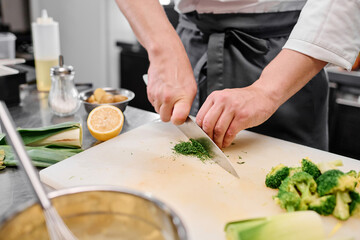Obraz na płótnie Canvas Close-up of chef using kitchen knife to cut broccoli in little pieces for dish on cutting board at table