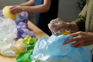 Hands of contemporary senior woman putting smashed plastic bottle into sack with similar waste while sorting trash with her grandson