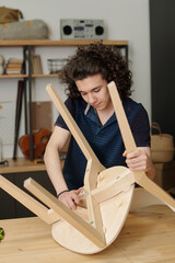 Modern male teenager in casualwear bending over table in garage while repairing wooden chair or making new one for his family