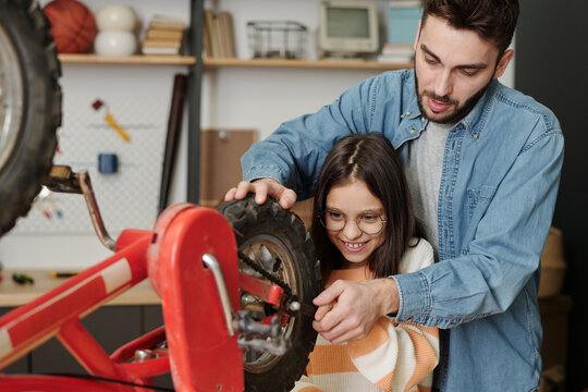 Smiling little girl helping father repair her bicycle while holding part of wheel and using wrench to fix it in garage with household supplies