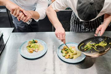Close-up of cook putting garnish from pan on plate with his colleague adding spices on ready dish, they working in kitchen