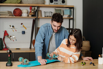 Cute little girl fixing wheels of blue skateboard with handtool while standing by wooden table in...