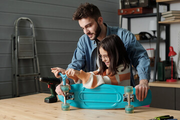 Cute smiling girl with handtool fixing wheels of skateboard on wooden table in garage while young...