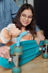 Fototapeta na wymiar Smiling cute youthful girl in eyeglasses fixing wheels of blue skateboard with wrench while standing by table with her father on background