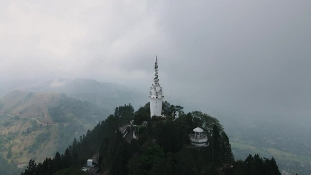 High white Ambuluwawa tower with the spiral stairs to the viewpoint on top of the Ambuluwawa mountain while the valley is partly covered with low hanging clouds. Wide backwards drone dolley shot