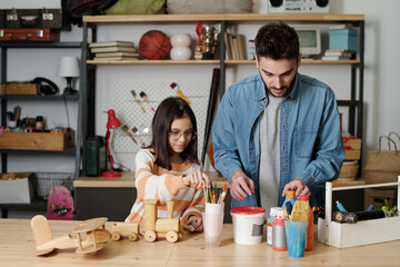 Young creative man and little girl in casualwear preparing gouache and paintbrushes while going to paint handmade wooden toys