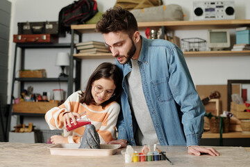 Cute little girl pouring red liquid into handmade volcano crater while standing by table in garage and making mockup with her father