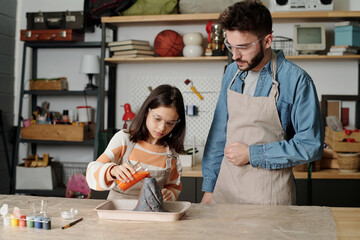 Father and daughter in aprons and protective eyeglasses pouring orange liquid into crater of...