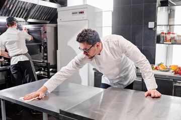 Chef in uniform wiping kitchen table after cooking with his colleague cleaning the oven after work...