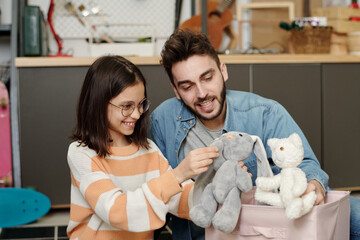 Happy little girl with grey soft toy rabbit and her father with white teddybear playing together in garage at leisure