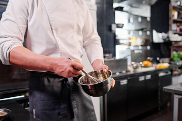 Close-up of chef in apron whipping eggs in bowl with a whisk while cooking in kitchen