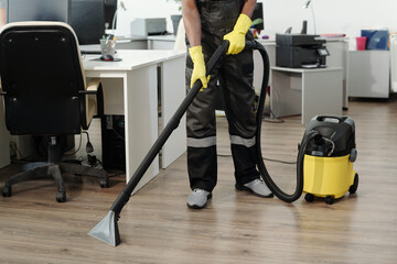 Young black man in uniform and yellow gloves using vacuum cleaner for cleaning floor of large...