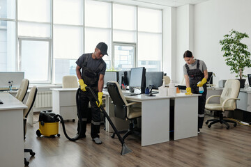Young black man in workwear cleaning floor while girl wiping desks with computer monitors in large contemporary openspace office